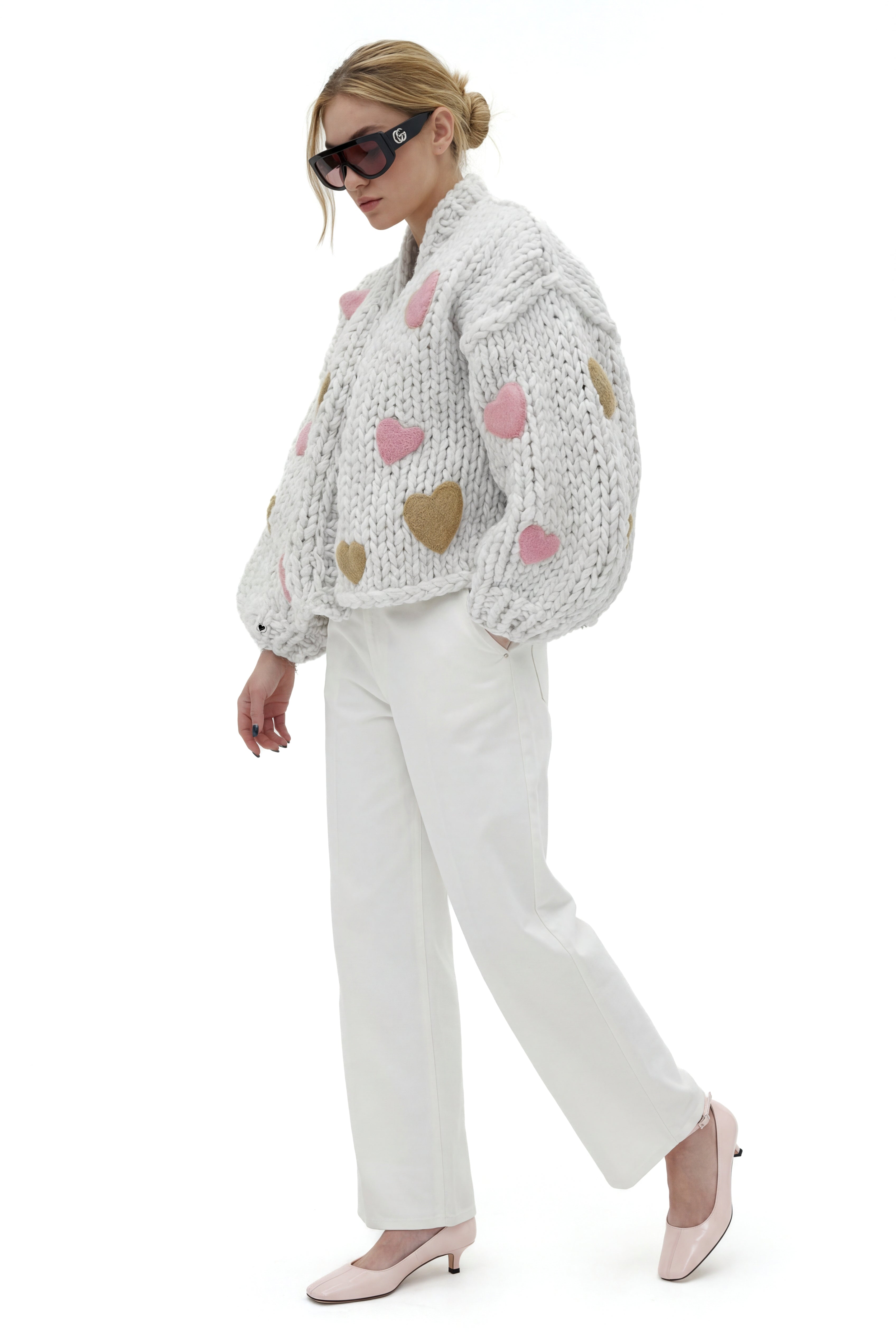 Woman wearing a knitted sweater with heart on and white pants on a white background